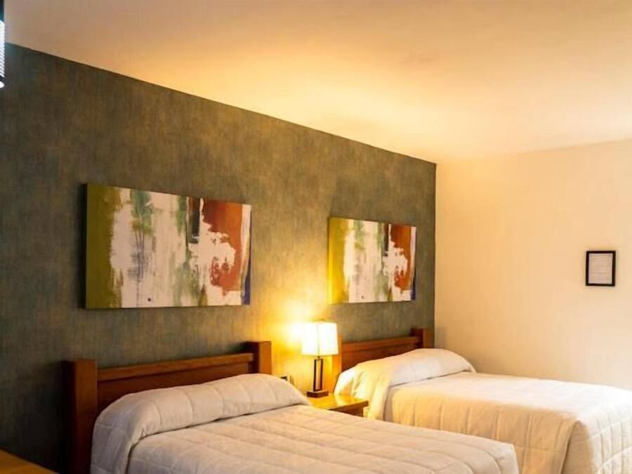 A hotel room with two beds, a nightstand with a lamp, and two abstract paintings on the wall.