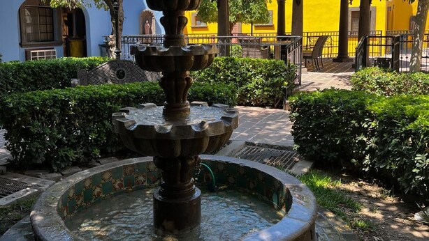 Ein Innenhof mit einem zentralen Brunnen, umgeben von Grünflächen und einem gelben Gebäude mit einem Ziegeldach.