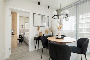 Ein modernes Hotelzimmer mit Holztür, einem runden Esstisch mit schwarzen Stühlen, einem großen Fenster mit leichtem Vorhang und einer Deckenleuchte.
