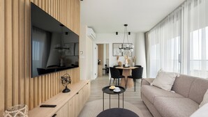 Een moderne woonkamer met een flatscreen televisie, een bank, een eethoek met stoelen en een groot raam met dunne gordijnen.