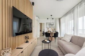 Un salon moderne doté d’un téléviseur à écran plat, d’un canapé, d’un coin repas avec des chaises et d’une grande fenêtre agrémentée de rideaux légers.