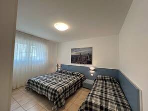 A room with two beds, a cityscape painting, a window with sheer curtains, and a ceiling light. - Modern Flat 50mt From the Beach (Lignano Sabbiadoro)