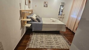 A narrow room with a bed, a small shelf, a patterned rug, and a mirror.