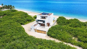 A modern two-story beachfront house with a flat roof, large windows, and a visible solar panel array.