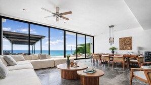 A modern living room with a large window overlooking the ocean, a ceiling fan, and a dining area with wooden furniture.