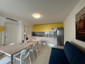 A modern kitchen with yellow cabinets, a stainless steel refrigerator, and a dining area with a wooden table and white chairs. - Modern Flat With Balcony by the Beach (Lignano Sabbiadoro)