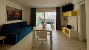 A modern living room with a sofa, dining table, chairs, a TV mounted on the wall, and a balcony with a view of buildings. - Modern Flat With Balcony by the Beach (Lignano Sabbiadoro)
