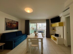 Ein modernes Wohnzimmer mit Sofa, Esstisch, Stühlen, einem an der Wand befestigten Fernseher und einem Balkon mit Blick auf Gebäude.