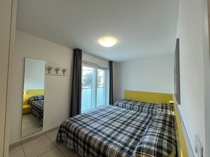 A hotel room with two beds, a large mirror, a window with curtains, and a ceiling light. - Modern Flat With Balcony by the Beach (Lignano Sabbiadoro)