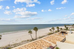 A beach view from a balcony with palm trees, a pool area, and a beach with people. - Newly Remodeled 2/2 Gulf Front Condo in Madeira Beach, Florida (Madeira Beach)