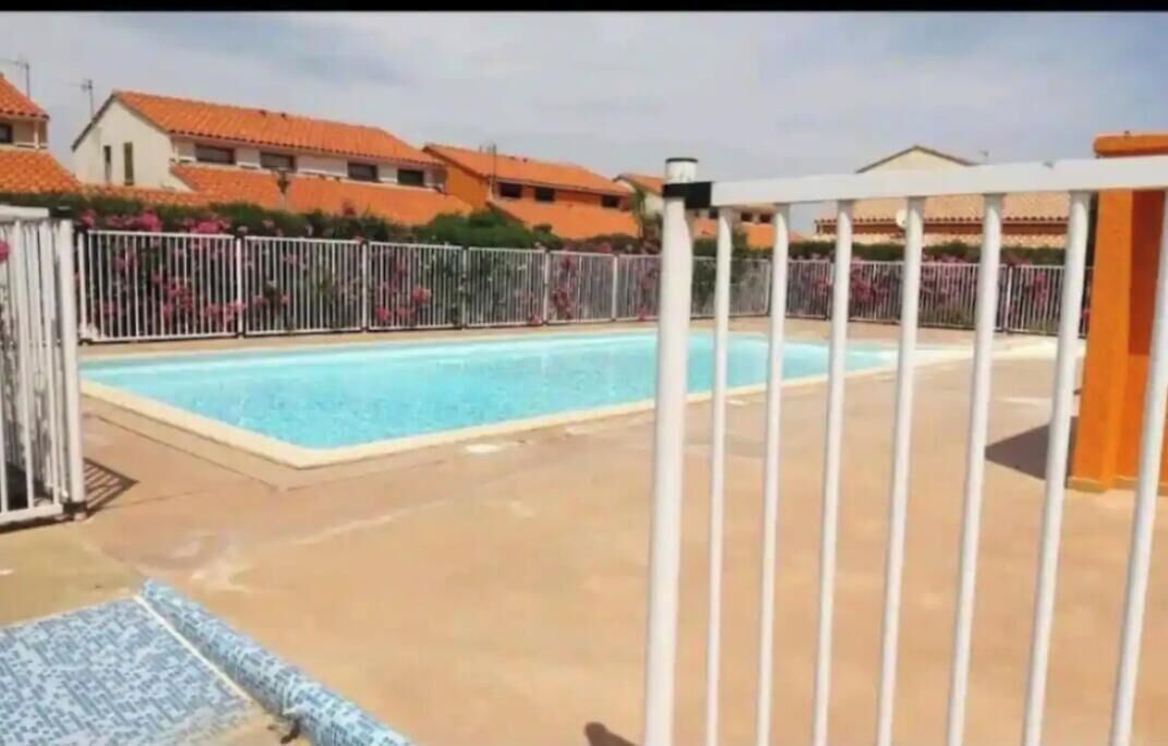 Una piscina con valla blanca, zona cubierta y edificios al fondo.