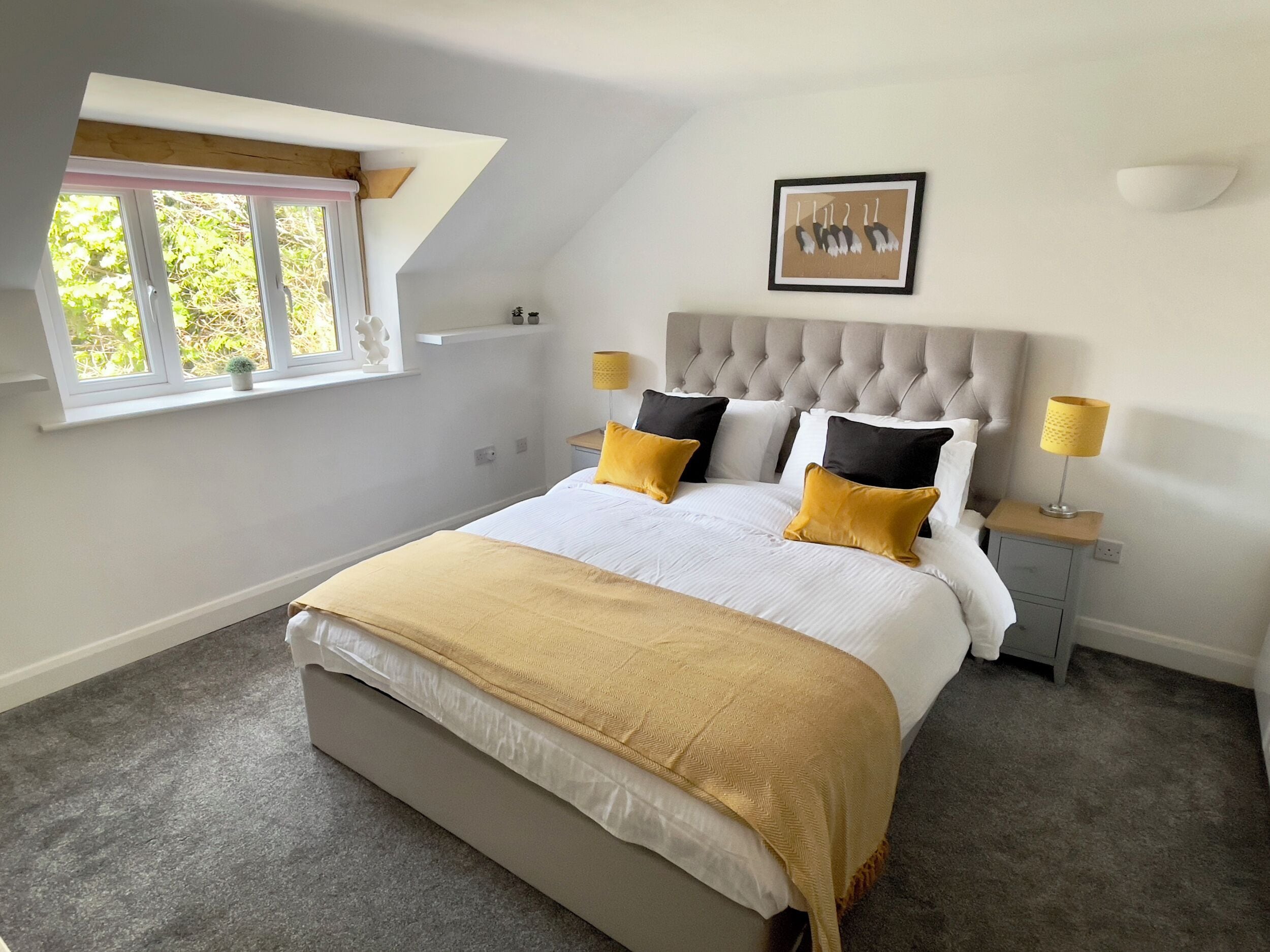 A bedroom with a large bed, a window with a view, a headboard, bedside tables, and a picture on the wall.