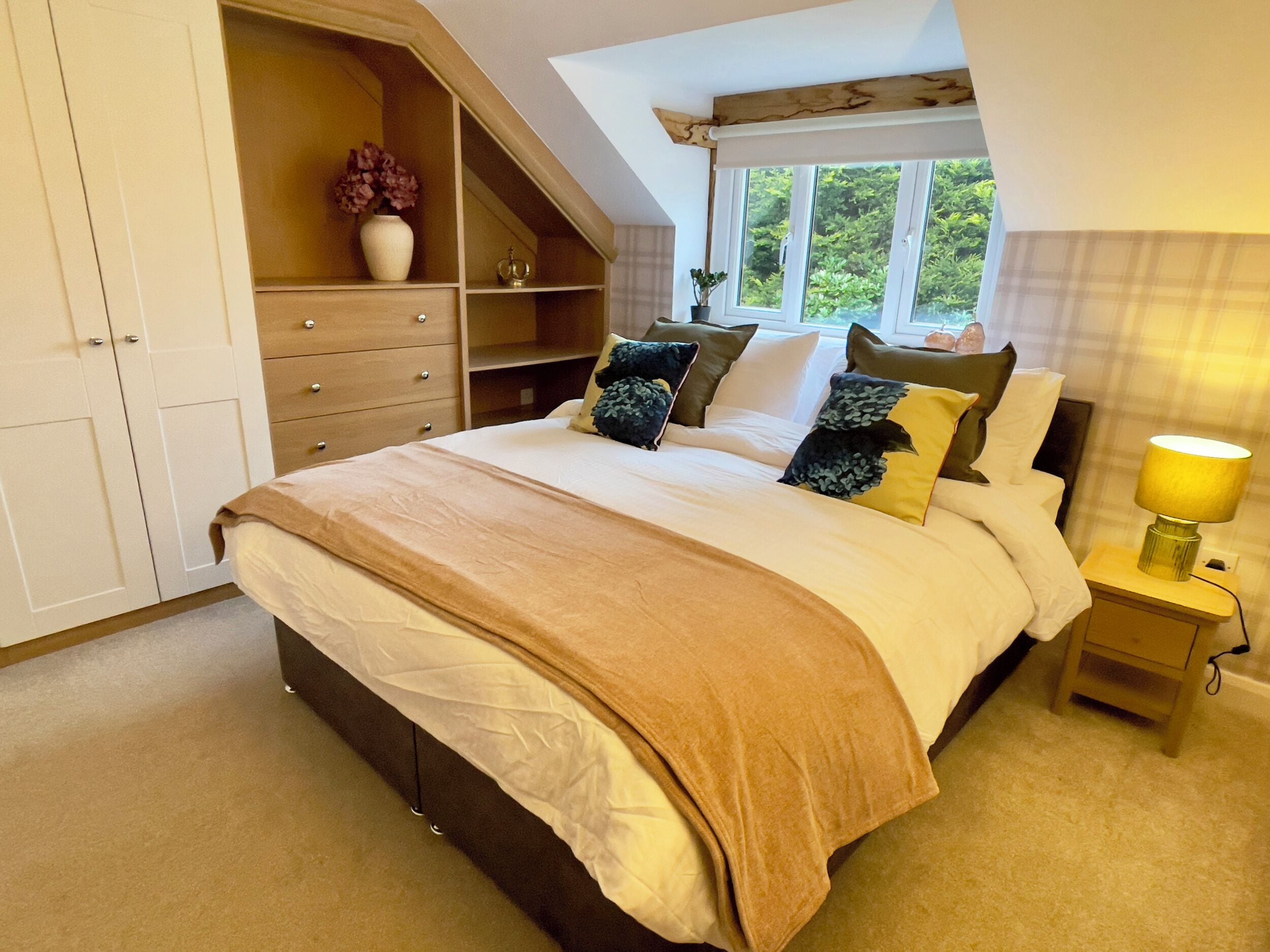 A bedroom with a large bed, a nightstand with a lamp, a wardrobe, and a window with a view of greenery.