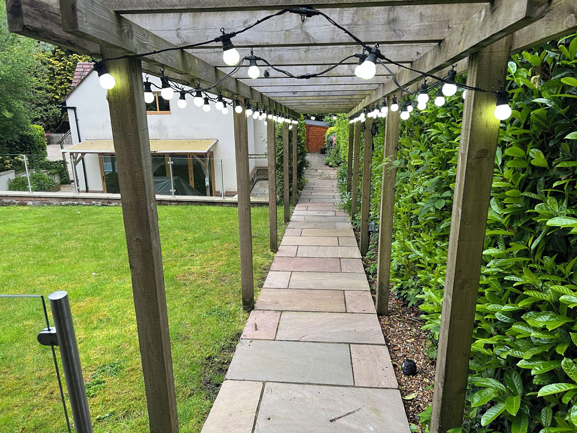 A stone pathway under a wooden pergola with string lights, surrounded by greenery and a house in the background.