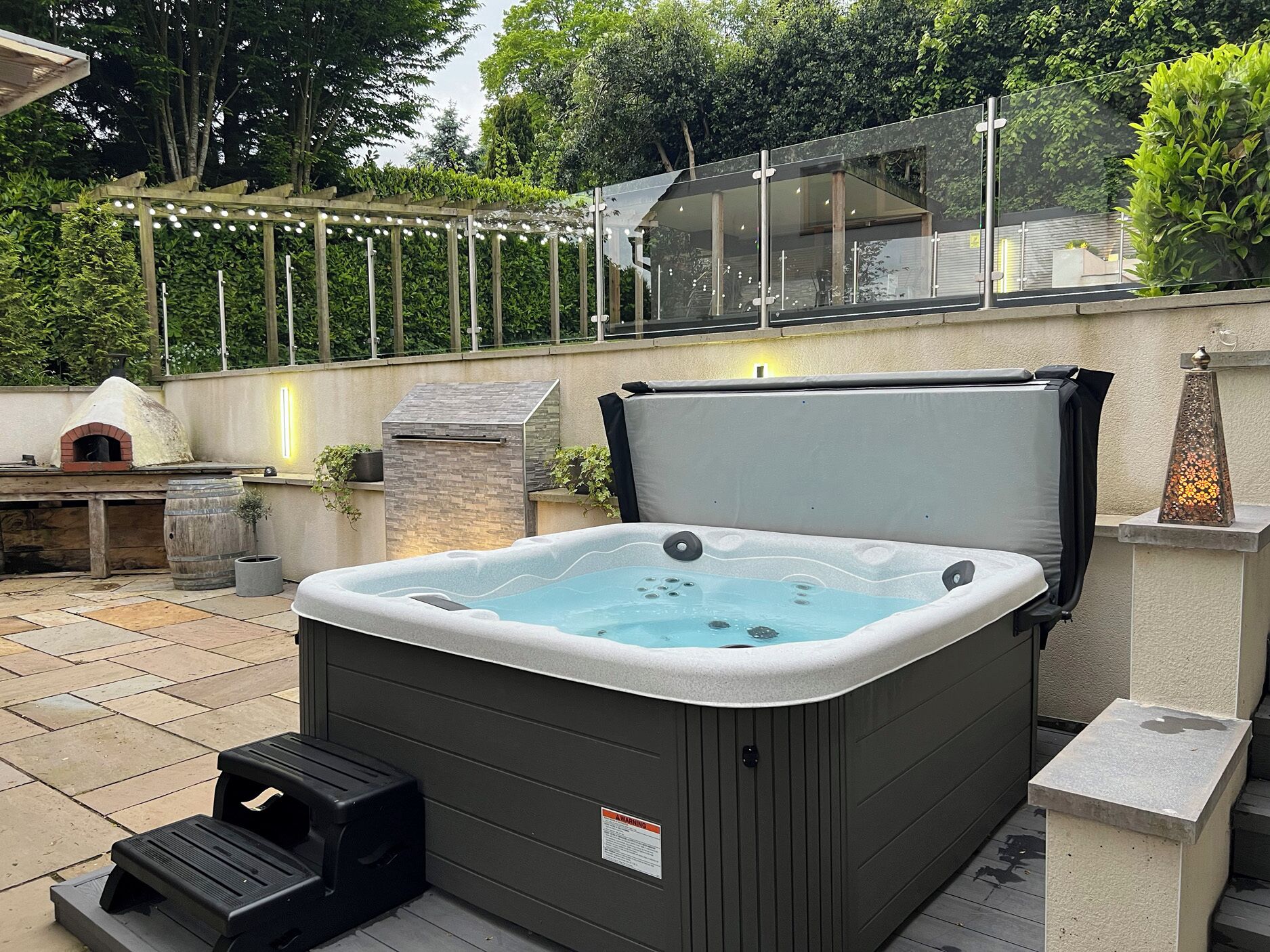 A hot tub with a stone surround, a wooden pergola with string lights, and a stone fireplace.