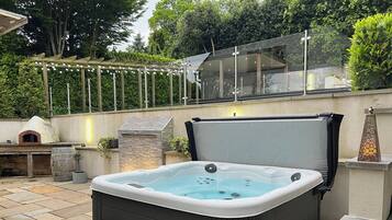 A hot tub with a stone surround, a wooden pergola with string lights, and a stone fireplace.