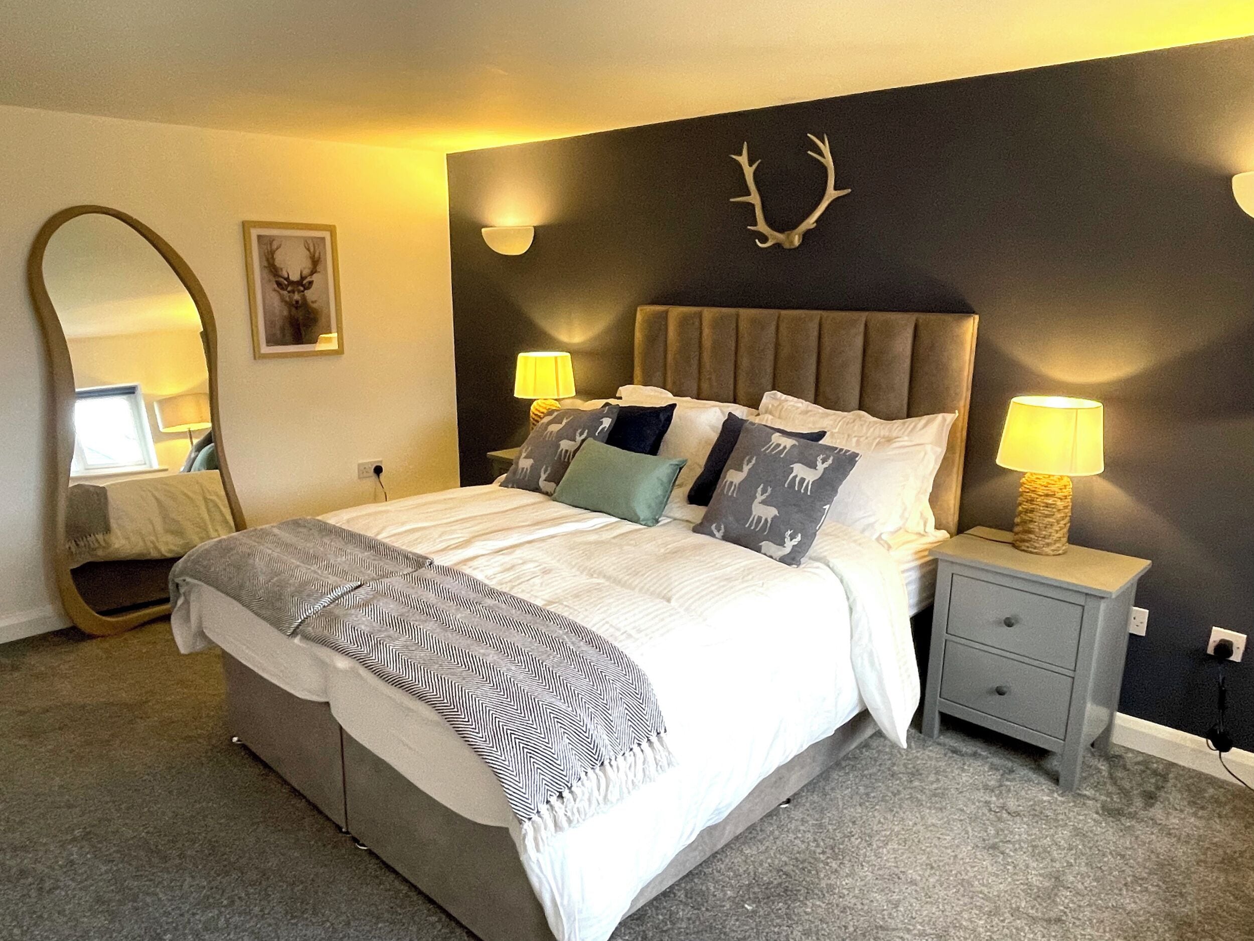 Un dormitorio con una cama, dos mesitas de noche, un espejo, una decoración de cabeza de venado montada en la pared y una lámpara en cada mesita de noche.