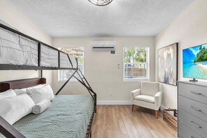 A hotel room with a bunk bed, a single bed, a chair, a TV, and an air conditioning unit. - Sunset Dreams (St. Pete Beach)