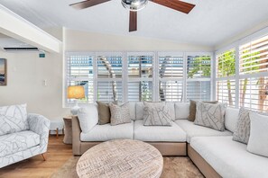 A modern living room with a sectional sofa, a round coffee table, and large windows with blinds. - Sunset Dreams (St. Pete Beach)