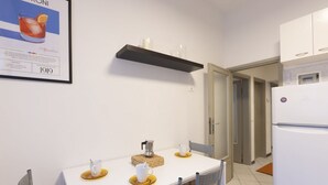 Eine kleine Küche mit einem weißen Tisch, zwei Stühlen, einem schwarzen Regal und einem weißen Kühlschrank.