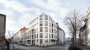 A modern multi-story building with a curved facade, a pedestrian crossing, and parked cars along the street. - Just Sunny – 2.5-Room Apt near Messe & City Center (Basel)