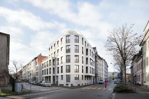Un bâtiment moderne à plusieurs étages, doté d’une façade incurvée, d’un passage piéton et de voitures garées le long de la rue.