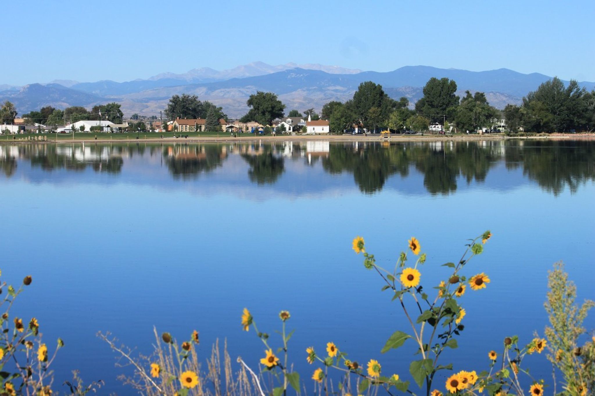 Un lac paisible où se reflètent les arbres et les montagnes, entouré d’une petite ville avec ses maisons et un ciel dégagé.