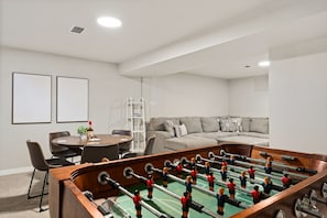 Een kamer met een tafelvoetbaltafel, een bank, een eettafel met stoelen en een kleine plant.