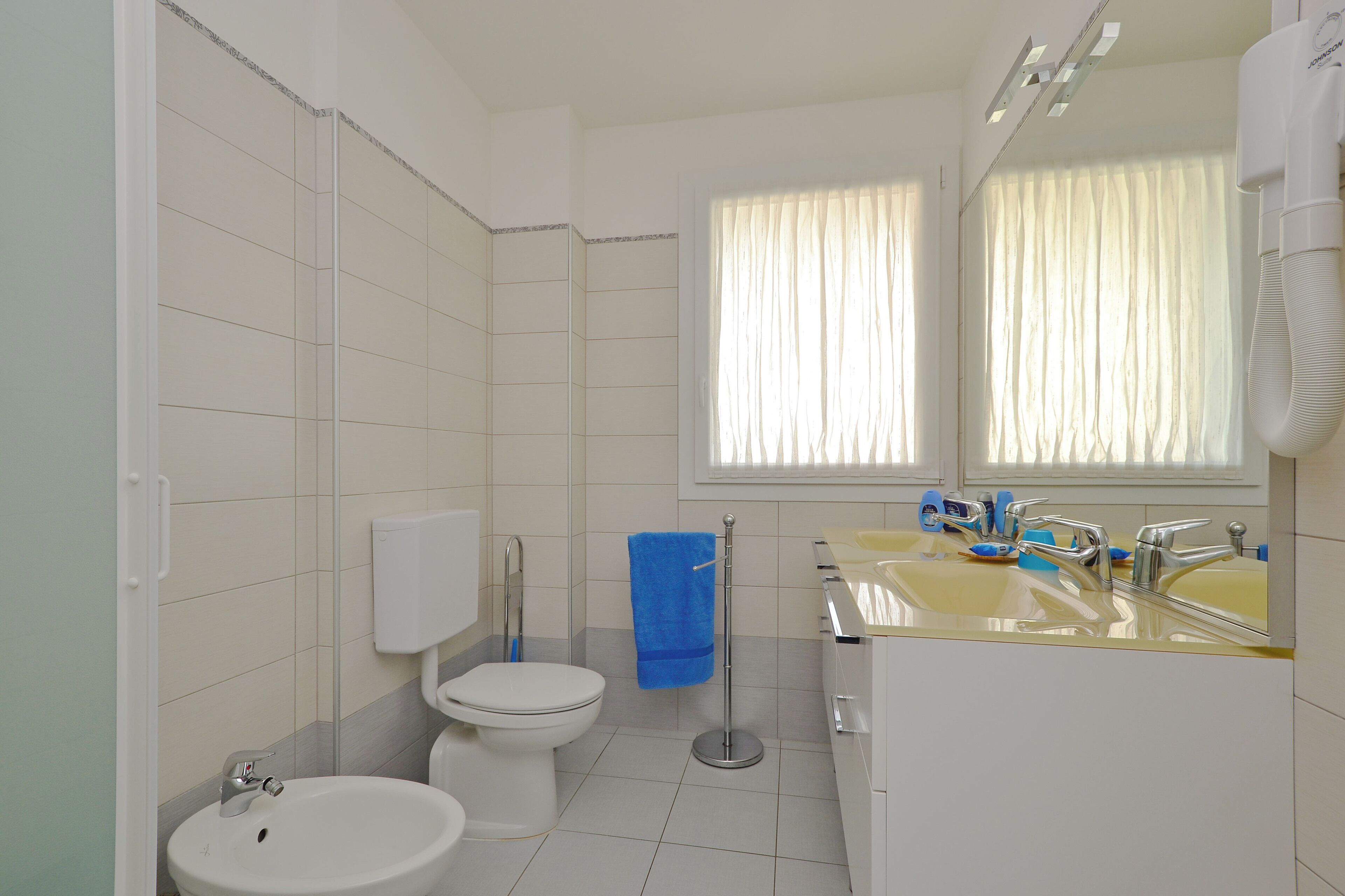 A bathroom with a toilet, bidet, sink, and a window with curtains.