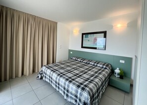 A bedroom with a bed, curtains, a wall-mounted TV, and a small bedside table with a plant. - Penthouse With Rooftop Terrace and sea Views (Lignano Sabbiadoro)