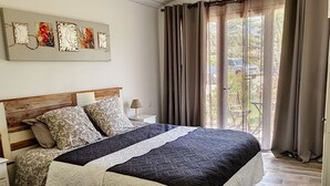 Un dormitorio con cabecera de madera, una cama con ropa de cama estampada, una mesita de noche con lámpara y vistas al exterior a través de una ventana con cortinas.