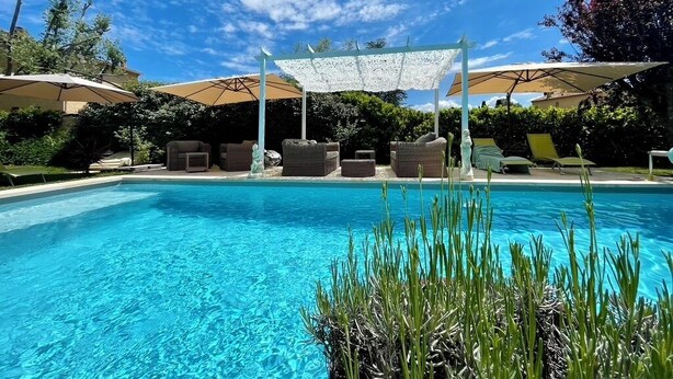 Piscina con sillones de descanso y sombrillas, rodeada de follaje.