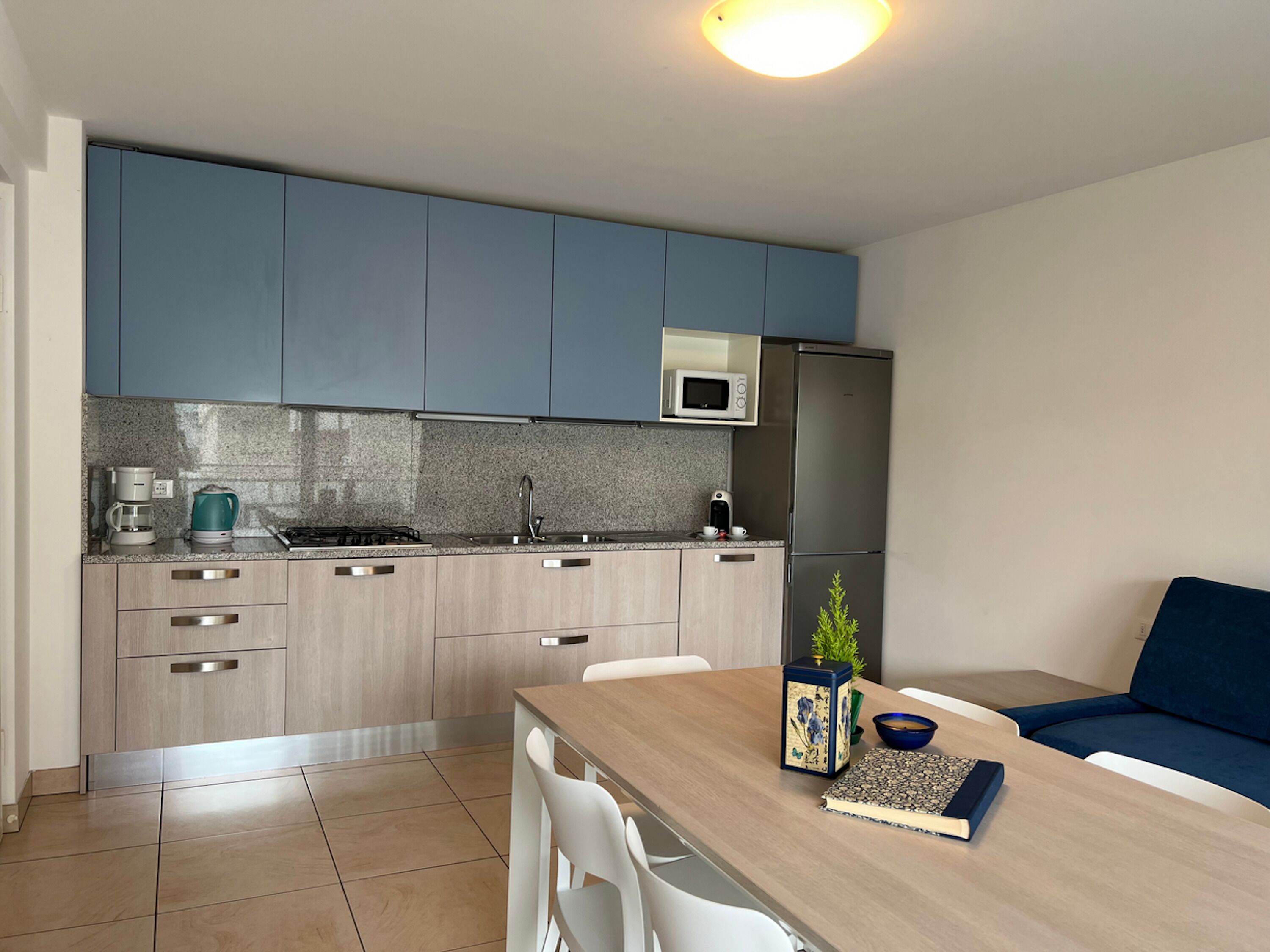 A modern kitchen with grey cabinets, a microwave, and a refrigerator. There is a dining table with chairs and a blue sofa.