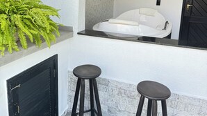 Uma área de estar moderna ao ar livre, com um sofá branco e dois banquinhos, uma porta preta e uma pintura decorativa de um cavalo na parede.