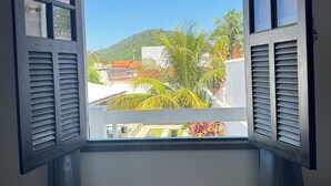 Quarto com janela, persianas de madeira, uma cama e vista para uma colina verde e edifícios.