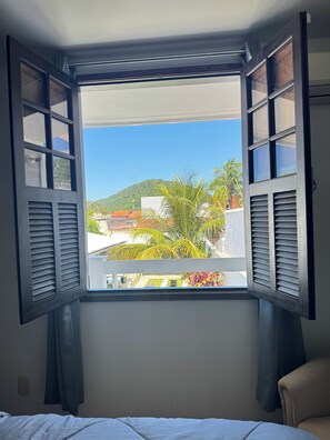 A room with a window featuring wooden shutters, a bed, and a view of a green hill and buildings. - Spacious vacation home for 10 people with pool 200 meters to the beach (Niterói)