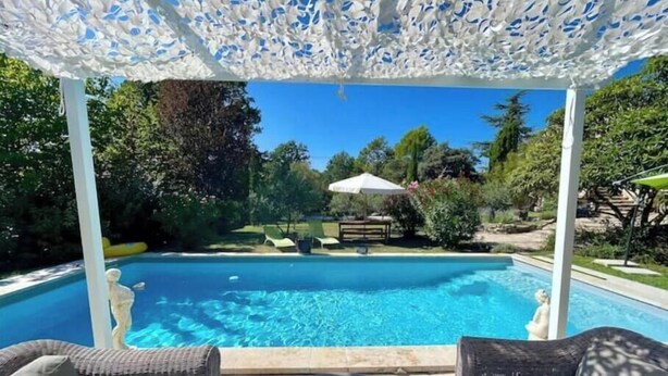 A swimming pool with a white pergola overhead, surrounded by greenery and outdoor furniture. - La belle histoire du gîte Ventoux (Bédoin)