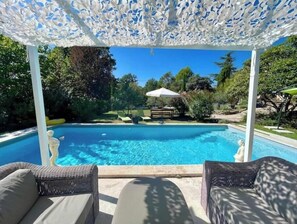 Una piscina con una pérgola blanca sobre ella, rodeada de vegetación y mobiliario de exterior.