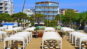 A beachfront hotel with white canopied seating areas, a blue and white building in the background, and a flagpole with a flag. - Central Lignano Flat With Balcony (Lignano Sabbiadoro)
