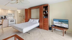 Un dormitorio con cama, televisor, ventilador, kitchenette y un mueble de madera.