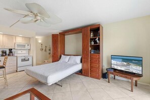 Ein Schlafzimmer mit Bett, Fernseher, Ventilator, einer Küchenzeile und einem Holzschrank.