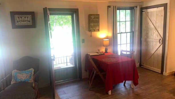 Una stanza con pavimento in legno, un tavolo rosso, una sedia con cuscino, una porta e una finestra.