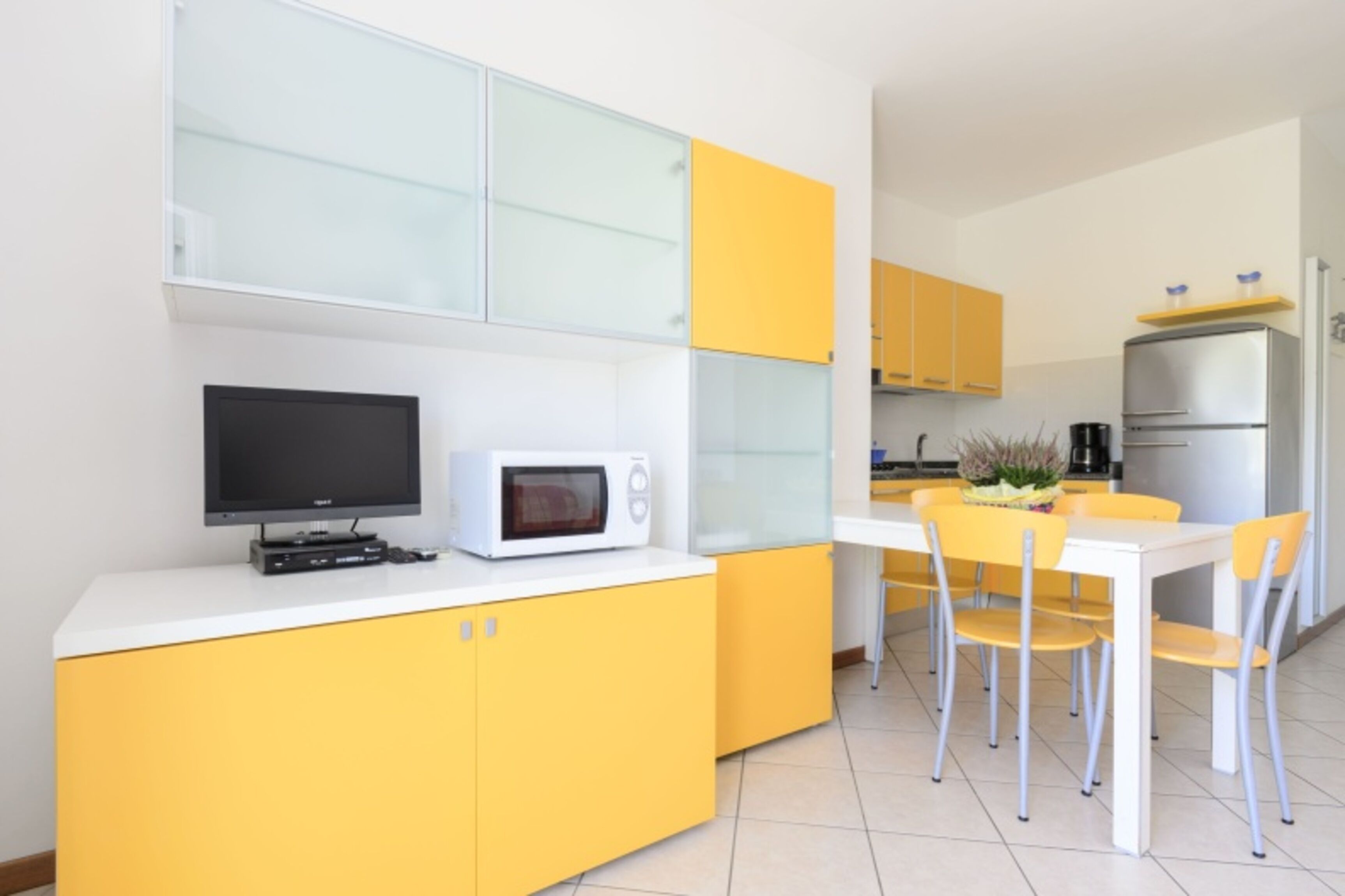 Une cuisine moderne avec des meubles de couleur jaune, un plan de travail blanc, un four à micro-ondes, une télévision et un coin repas avec des chaises jaunes.