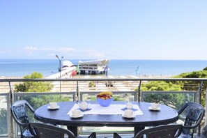Een ronde tafel met een schaal fruit, witte kopjes en een theepot op een balkon met uitzicht op zee.