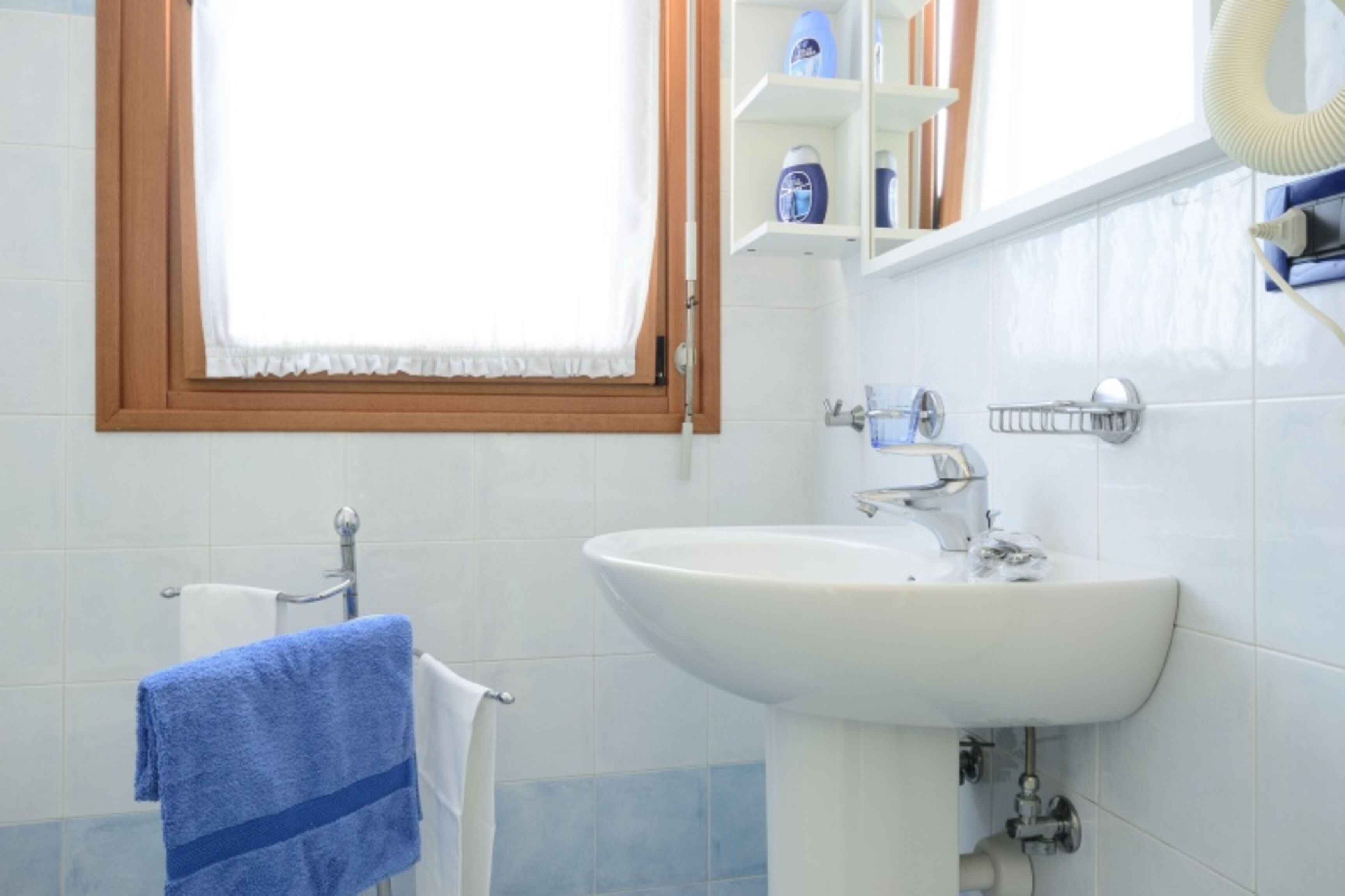Un bagno con un lavandino bianco, un porta asciugamani, una finestra con cornice in legno e uno scaffale con articoli da toilette.