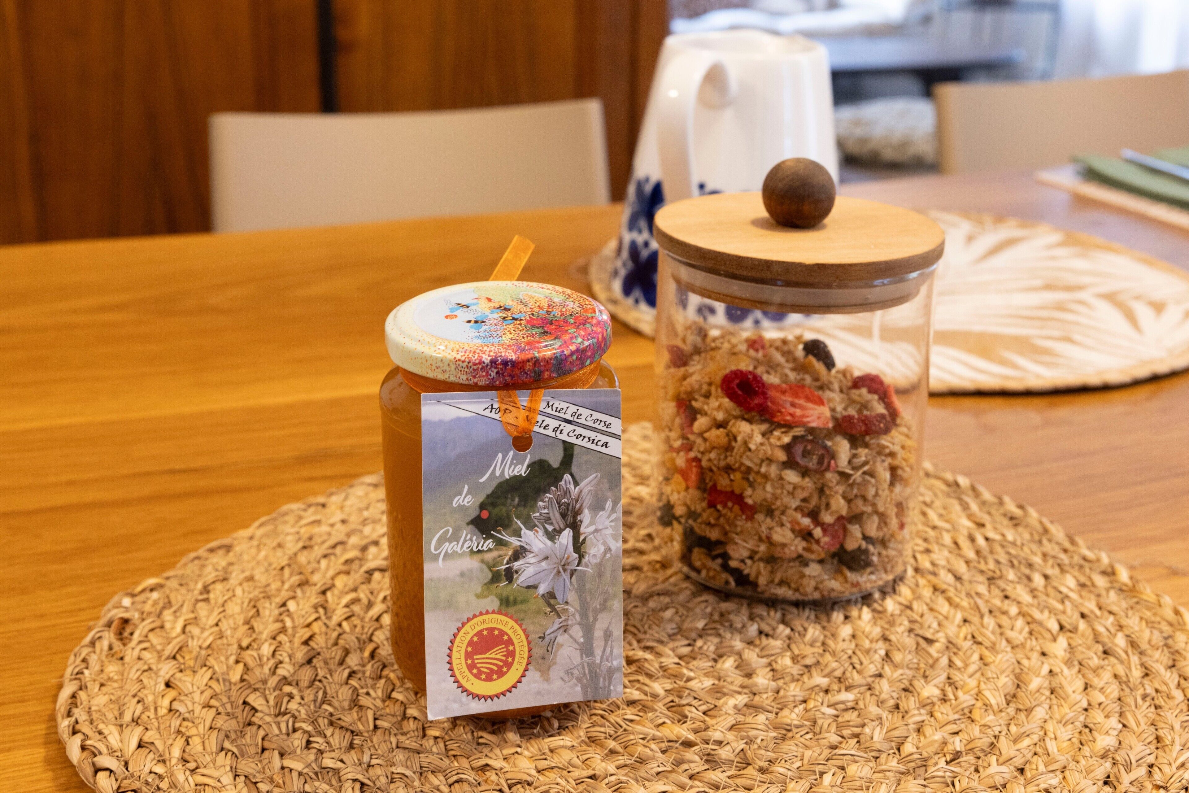 Um pote de granola com tampa de madeira e etiqueta, sobre um descanso de palha em uma mesa de madeira.