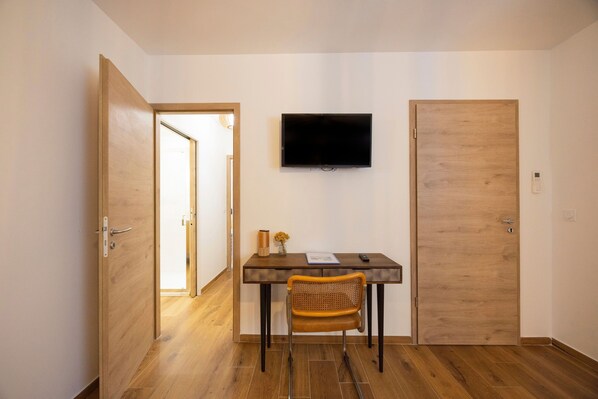 A room with a wooden floor, a desk with a chair, a flat-screen TV mounted on the wall, and a closed door.