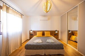 A hotel room with a bed, bedside lamps, a ceiling light fixture, a window with curtains, and a wall-mounted air conditioning unit.