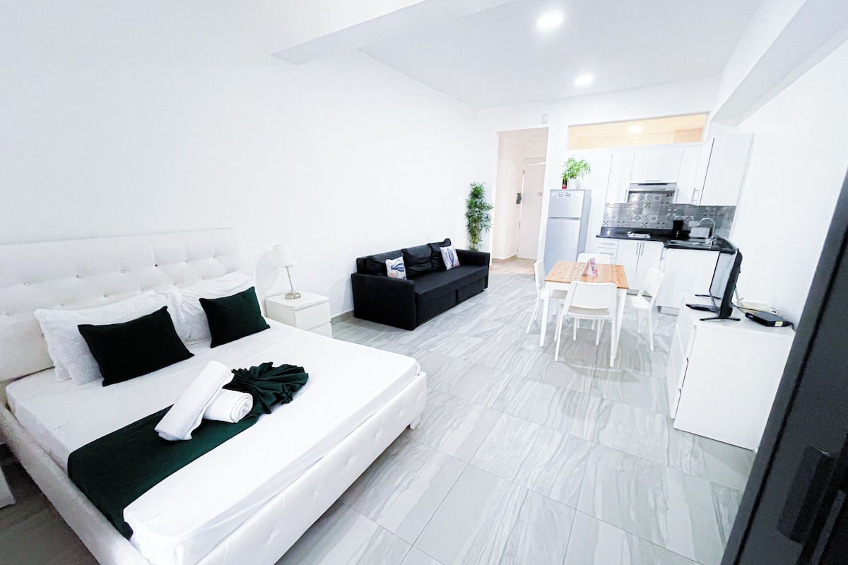 A modern hotel room with a large bed, a black sofa, a dining table with chairs, a kitchen area, and a small plant.