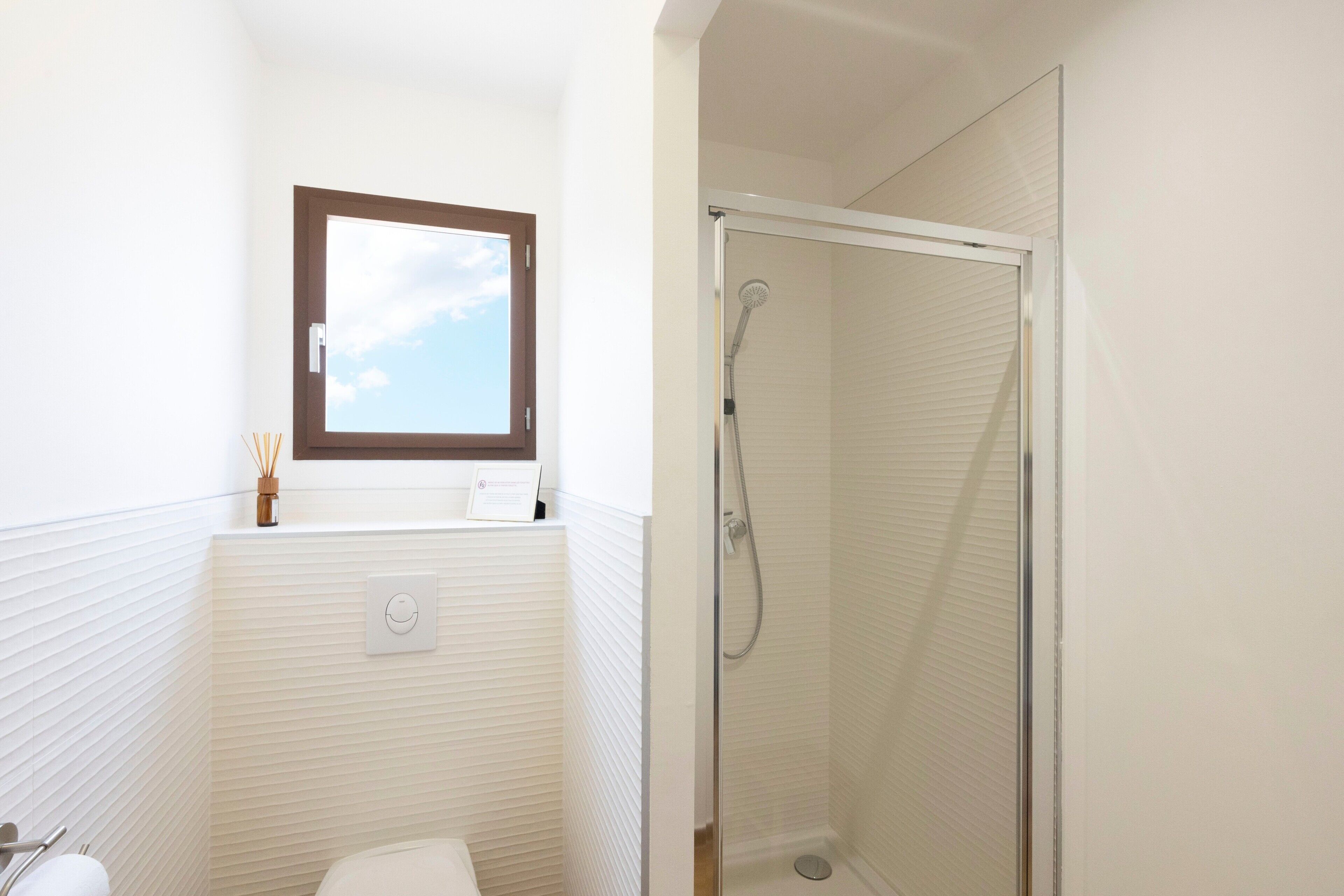 Een badkamer met toilet, douche, raam en een lichtschakelaar aan de muur.